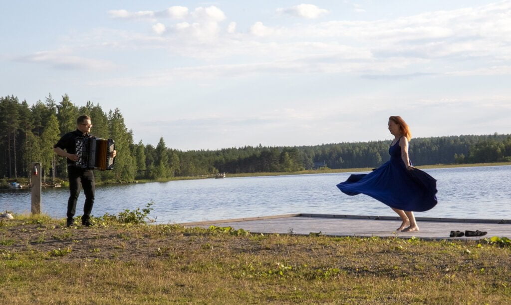 Nainen tanssii laiturilla, mies soittaa hanuria.