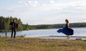 Nainen tanssii laiturilla, mies soittaa hanuria.